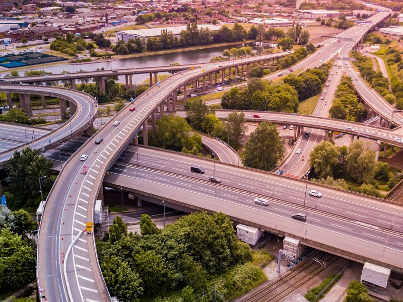 Aerial view of a complex motorway road junction with traffic moving. Cars, lorries, vans and a train can be seen travelling through a busy road interchange from above.
