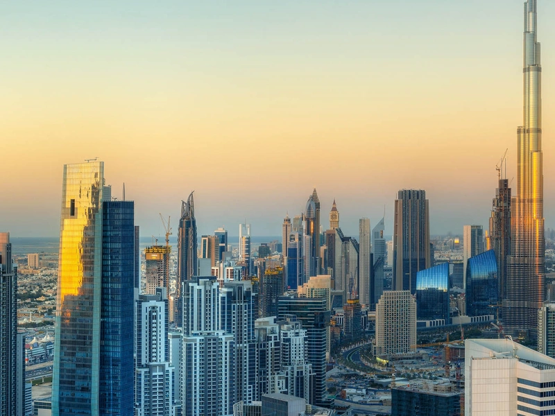 A panoramic view of a modern city skyline at sunset, featuring numerous high-rise buildings and skyscrapers, including the iconic Burj Khalifa on the right, dominating the landscape with its towering height. The buildings are illuminated by the warm golden light of the setting sun, creating a striking contrast against the clear sky.