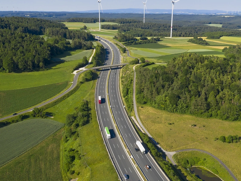 Traffic on a motorway surrounded by green fields, forest and wind turbines in the far distance