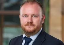 David Griffiths, Director, Real Estate Lead, UAE,  outside in front of a blurred brown background featuring a building and mix of natural and urban elements.