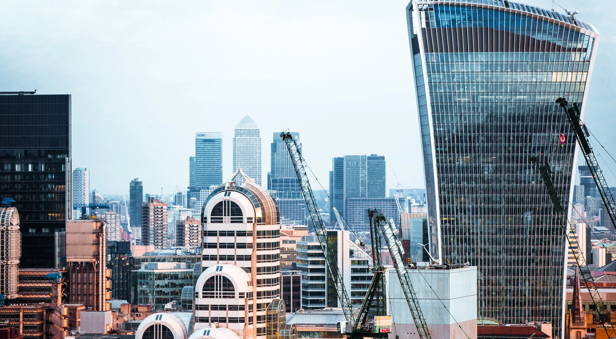 A view of the London skyline at dusk, bathed in a pinkish-golden light, with a cloudy sky. Four construction cranes are visible, hinting at ongoing construction work in the area.