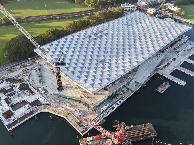 Ariel view of a large waterfront construction site with a geometric roof structure and surrounding cranes and platforms