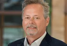 Portrait of Ron Staa, Director, outside in front of a blurred brown background featuring a building and mix of natural and urban elements.