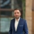 Paul Barry, outside in front of a blurred brown background featuring a building and mix of natural and urban elements.