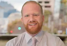Portrait of Chris Robinson, Associate Director, Contract Services, outside in front of a blurred brown background featuring a building and mix of natural and urban elements.